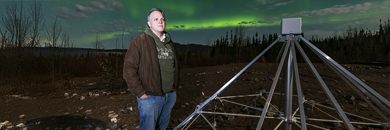Jonathan Sievers debout à côté d’une antenne radio à basse fréquence située à la station Uapishka, près du réservoir Manicouagan, dans le nord du Québec. Les aurores en arrière-plan permettent à l’équipe d’étudier l’impact de l’ionosphère terrestre sur les données de radioastronomie. © Babak Tafreshi